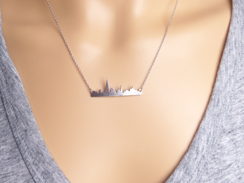 Puede incluir: Un collar de plata con un colgante de ciudad. El colgante es una silueta detallada de un horizonte urbano.