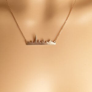 May include: A gold necklace featuring a delicate cityscape pendant. The pendant is a silhouette of a city skyline with tall buildings.