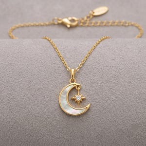 May include: A gold-tone necklace featuring a crescent moon pendant inlaid with iridescent white mother-of-pearl. A small, four-pointed star charm set with a clear, sparkling stone hangs within the curve of the moon. The chain includes a lobster clasp.