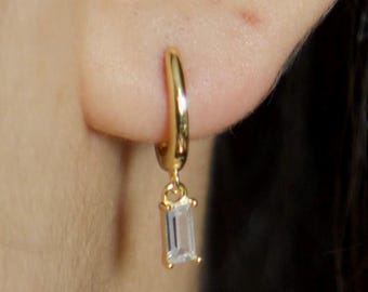 Puede incluir: Aretes de aro dorados con una piedra preciosa rectangular transparente colgante.