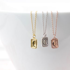 May include: Three delicate necklaces with rectangular pendants featuring a crescent moon and sunburst design. The necklaces are gold, silver, and rose gold, each with a fine chain. The pendants are approximately 0.5 inches in height.