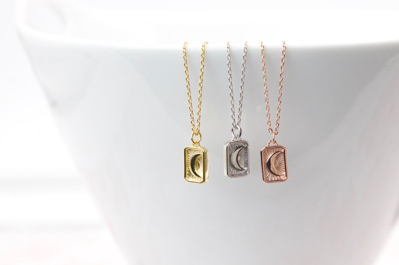 May include: Three delicate necklaces with rectangular pendants featuring a crescent moon and sunburst design. The necklaces are gold, silver, and rose gold, each with a fine chain. The pendants are approximately 0.5 inches in height.