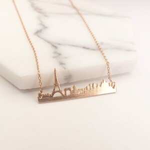 May include: A gold necklace with a delicate chain and a pendant featuring a cutout silhouette of the Paris skyline, including the Eiffel Tower.