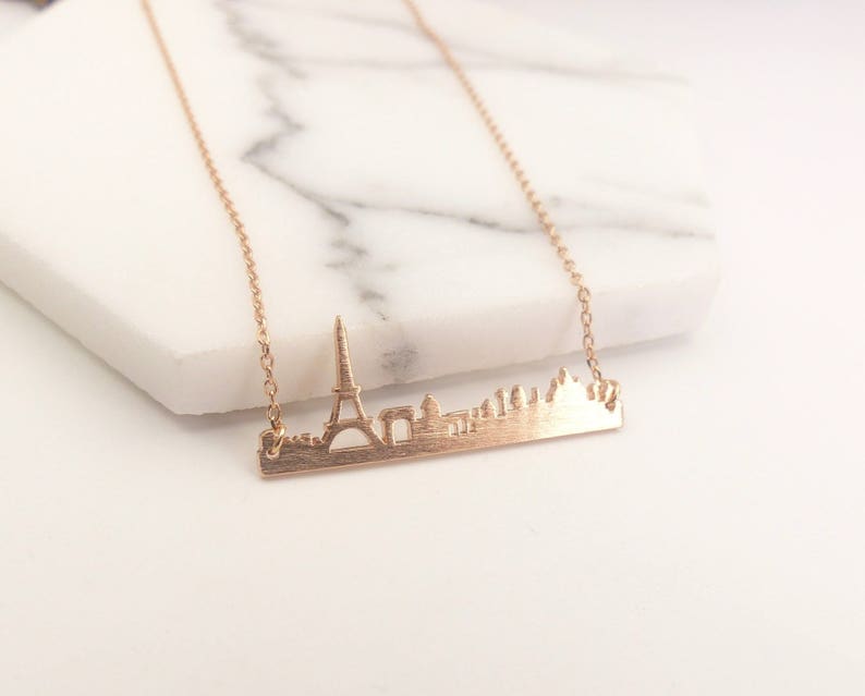 May include: A gold necklace with a delicate chain and a pendant featuring a cutout silhouette of the Paris skyline, including the Eiffel Tower.