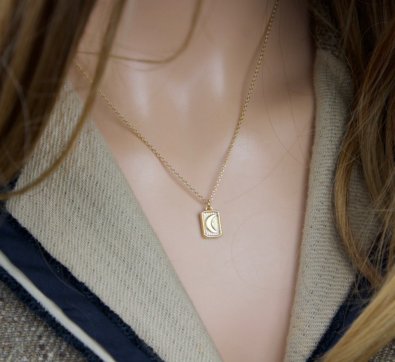 May include: A gold-colored necklace featuring a rectangular pendant with a crescent moon design. The pendant is suspended from a delicate chain. The necklace is displayed against a neutral background, highlighting the jewelry's details.