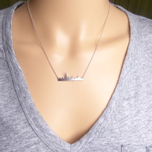 Puede incluir: Un collar de plata con un colgante de ciudad. El colgante es una silueta detallada de un horizonte urbano.