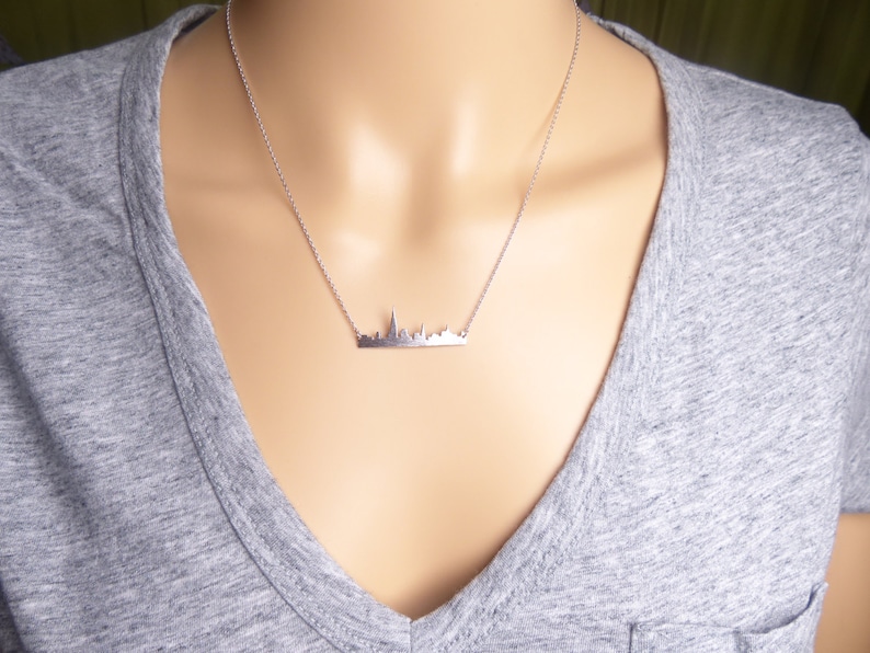 Puede incluir: Un collar de plata con un colgante de ciudad. El colgante es una silueta detallada de un horizonte urbano.