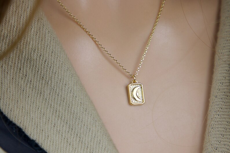 May include: A gold-colored necklace featuring a rectangular pendant with a crescent moon and sunburst design. The pendant hangs from a delicate chain. The necklace is worn against a neutral-toned background.