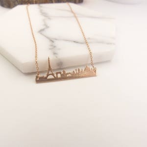 May include: A gold necklace with a delicate chain and a pendant featuring a detailed silhouette of the Paris skyline, including the Eiffel Tower.