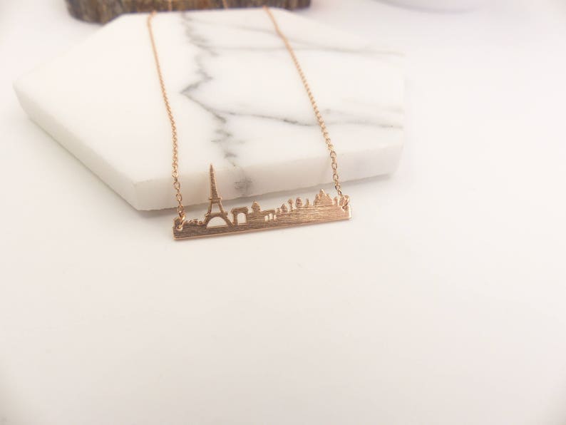 May include: A gold necklace with a delicate chain and a pendant featuring a detailed silhouette of the Paris skyline, including the Eiffel Tower.