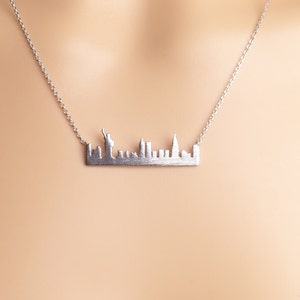 May include: A silver necklace featuring a New York City skyline pendant. The pendant is a detailed silhouette of the city's skyline, including the Statue of Liberty.