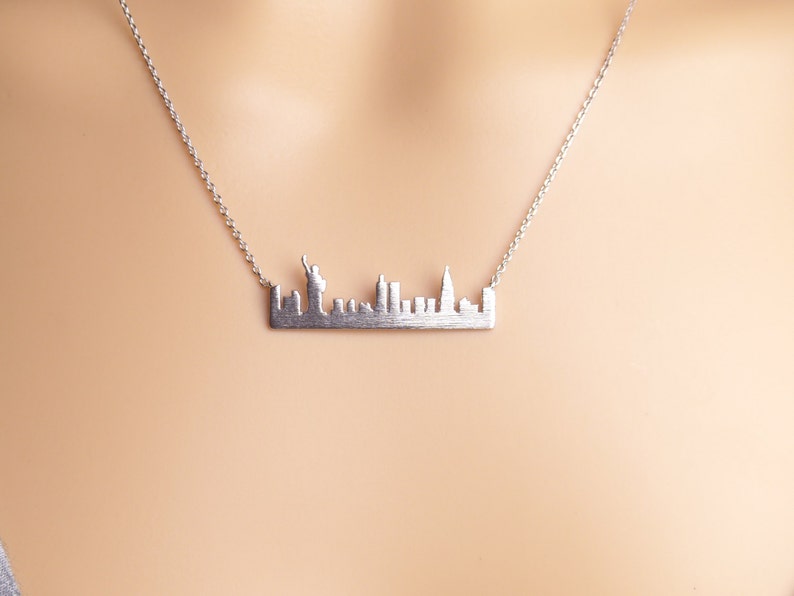 May include: A silver necklace featuring a New York City skyline pendant. The pendant is a detailed silhouette of the city's skyline, including the Statue of Liberty.