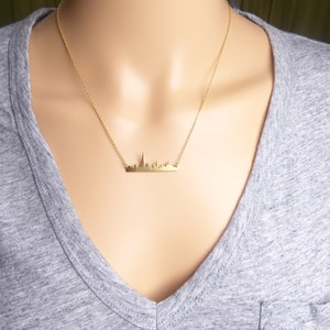 Puede incluir: Un collar de oro con un delicado colgante de ciudad. El colgante es una silueta simple de un horizonte urbano.