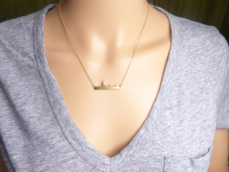 Puede incluir: Un collar de oro con un delicado colgante de ciudad. El colgante es una silueta simple de un horizonte urbano.