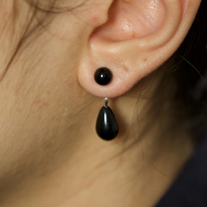 May include: A black, teardrop-shaped earring with a small, round stud. The earring is made of a glossy, dark material and hangs from a silver-colored post. The earring is worn on the ear of a person.