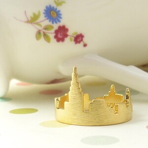 Puede incluir: Un anillo dorado con una silueta detallada de un horizonte de ciudad. El anillo est&aacute; sobre una superficie blanca con lunares de colores.