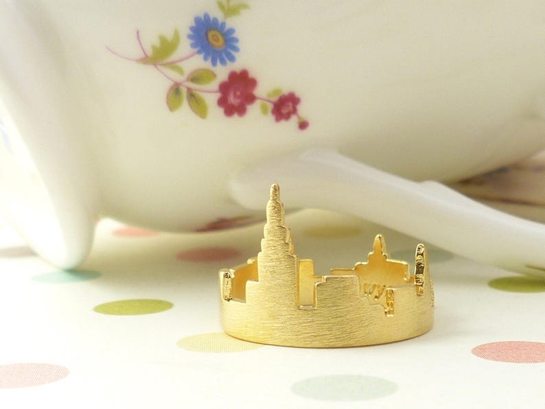 Puede incluir: Un anillo dorado con una silueta detallada de un horizonte de ciudad. El anillo est&aacute; sobre una superficie blanca con lunares de colores.