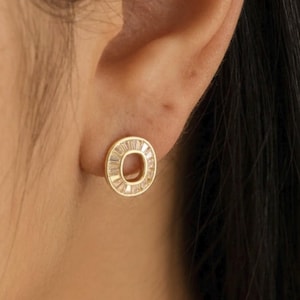 May include: A gold-colored stud earring with a circular design. The earring is adorned with small, clear, rectangular-cut stones around the outer edge. The earring is shown on an ear, with dark hair in the background.