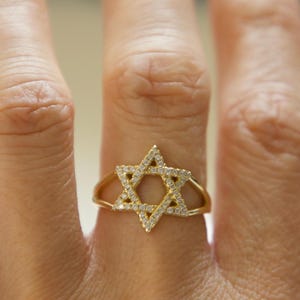 May include: A gold-colored ring featuring a Star of David design. The star is encrusted with small, clear gemstones. The ring is worn on a finger, showcasing the intricate details of the jewelry.