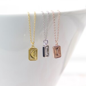 May include: Three delicate necklaces with rectangular pendants, each featuring a crescent moon design. The necklaces are gold, silver, and rose gold, suspended from fine chains. The pendants have a sunburst pattern around the moon.
