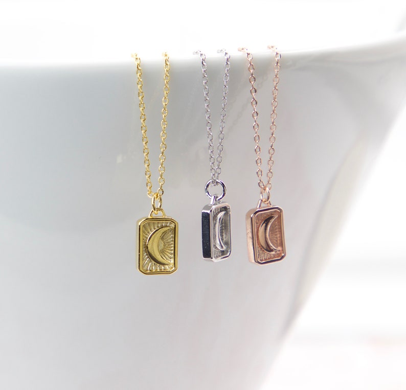 May include: Three delicate necklaces with rectangular pendants, each featuring a crescent moon design. The necklaces are gold, silver, and rose gold, suspended from fine chains. The pendants have a sunburst pattern around the moon.