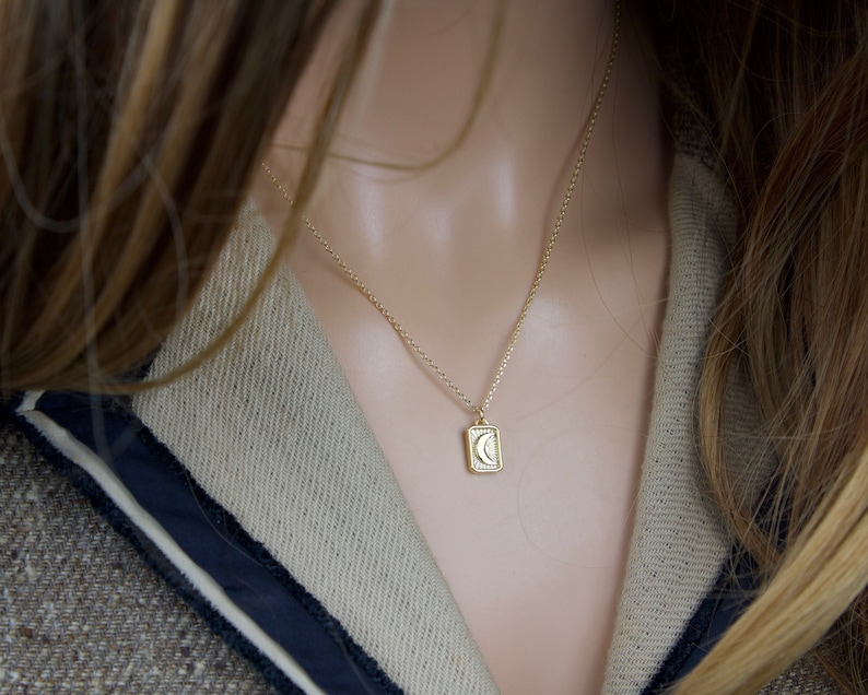 May include: A gold-colored necklace with a rectangular pendant featuring a crescent moon and sunburst design. The necklace is worn on a person with long, light brown hair and a tweed jacket with a navy blue trim.