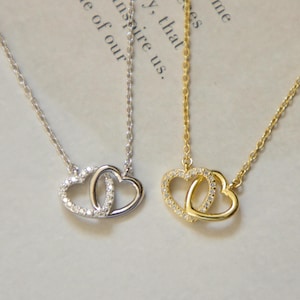 May include: Two necklaces featuring interlocking heart pendants. One is silver, the other gold. Both heart pendants are adorned with small, sparkling stones. The chains are fine and elegant, suitable for everyday wear. A romantic jewellery item.