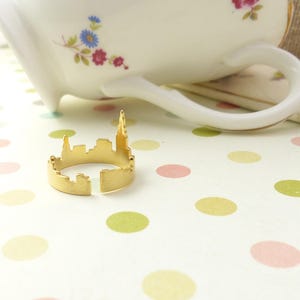 Puede incluir: Un anillo de color dorado con forma de horizonte de ciudad. El anillo est&aacute; sobre una superficie blanca con lunares rosas y verdes.