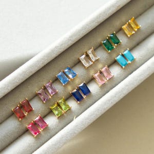 May include: A collection of gold-toned stud earrings, each featuring a rectangular gemstone in various colors. The gemstones include red, pink, purple, blue, green, yellow, and clear. The earrings are displayed in a gray jewelry tray.