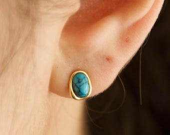 May include: A gold stud earring featuring an oval turquoise stone. The stone displays a marbled pattern with blue and green hues. The earring is set in a simple, polished gold bezel. The earring is shown on an ear.