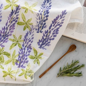 May include: A white tea towel with a floral pattern of purple lupine flowers and green leaves. The tea towel is folded and lying on a marble surface with a wooden spoon and a sprig of rosemary.