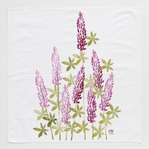 May include: A white tea towel with a floral design of purple lupine flowers and green leaves. The design is printed on the fabric.