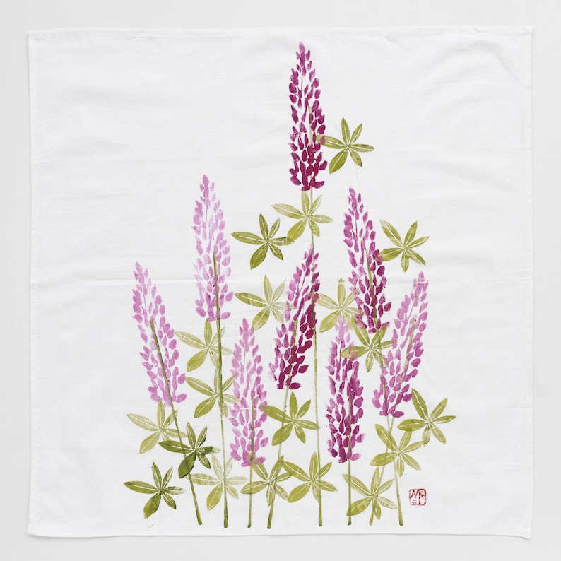 May include: A white tea towel with a floral design of purple lupine flowers and green leaves. The design is printed on the fabric.