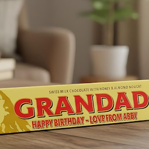 May include: A triangular yellow Swiss milk chocolate bar with honey and almond nougat. The packaging features a mountain graphic and the red text GRANDAD, followed by the text HAPPY BIRTHDAY - LOVE FROM ABBY in a smaller red font.