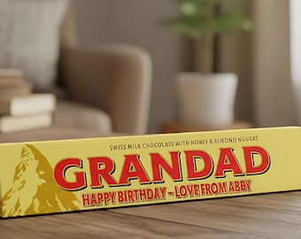 Può includere: Una barretta di cioccolato al latte svizzero triangolare con miele e torrone alle mandorle. La confezione presenta un grafico di montagna e il testo rosso GRANDAD, seguito dal testo HAPPY BIRTHDAY - LOVE FROM ABBY in un carattere rosso più piccolo.