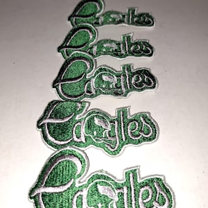 May include: Five embroidered patches with the word "Eagles" in green and white thread. The patches have a stylized eagle head design. The patches are arranged vertically on a white surface.