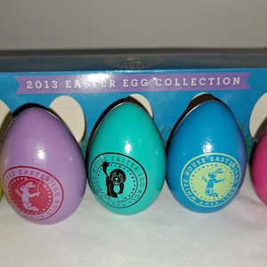 Puede incluir: Colección de cinco coloridos huevos de Pascua en amarillo, morado, turquesa, azul y rosa. Cada huevo tiene un diseño circular con un gráfico y el texto "WHITE HOUSE EASTER EGG ROLL 2013". Los huevos están frente a una caja azul.