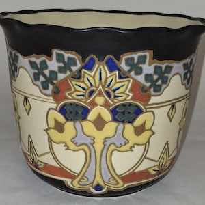 May include: A ceramic planter with a scalloped rim, featuring a cream-colored base decorated with a symmetrical pattern in shades of black, burnt orange, deep blue, and pale lavender. The design includes stylized floral and geometric motifs.