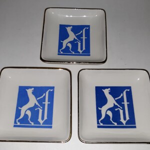 May include: Three square white ceramic dishes with a gold rim. Each dish features a blue square with a white dog silhouette and a stylized letter. The dishes are arranged in a triangular pattern.