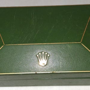 May include: A rectangular, green box with a gold trim and a gold emblem. The box has a textured surface and a hinged lid. The emblem is a gold crown. The box is likely for a watch or jewelry.