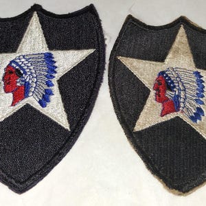 May include: Two embroidered patches featuring a black shield shape with a white star and a Native American profile. The profile is red with a blue and white headdress. The patches are likely military insignia.