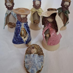 May include: A set of six ceramic nativity figurines featuring stylized figures with textured brown hair and beards. The collection includes five standing figures in colorful glazed cloaks and one infant figure lying in a textured brown ceramic manger.