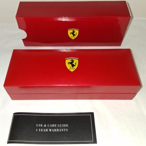 May include: Two red rectangular boxes with the Ferrari logo, one open. A black and white document with the text "USE & CARE GUIDE 1 YEAR WARRANTY" is below the boxes. The boxes are glossy and have a classic design.