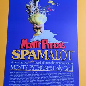 May include: A blue poster for the musical "Monty Python's Spamalot" features a golden chalice held by a hand, with the title in large yellow letters. The poster includes the text "A new musical lovingly ripped off from the motion picture Monty Python and the Holy Grail."
