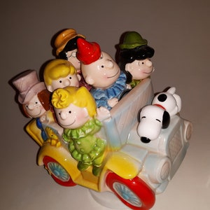 May include: A ceramic figurine featuring Peanuts characters in a yellow car with red wheels. The characters include Charlie Brown, Snoopy, and others, all with detailed facial features and colorful clothing. The car has a light blue body.