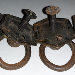 May include: Four antique metal drawer pulls with a dark, aged finish. Each pull features a square backplate with a decorative edge, a circular ring handle, and a screw for mounting. The metal has a rusty, weathered appearance.