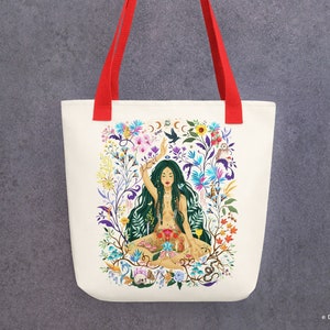 Op de afbeelding: Een witte tote bag met rode handvatten heeft een kleurrijke illustratie van een vrouw die in lotushouding zit, omringd door bloemen, planten en dieren. De vrouw heeft lang groen haar en draagt een jurk met bloemenprint. De tekst "DRAWMEASONG" staat onderaan de tas.