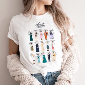 Può includere: T-shirt bianco con un'illustrazione colorata di donne della scienza storiche. Il design include il testo "Women Science" in un carattere corsivo, con i nomi delle donne sotto i loro ritratti. La maglietta è realizzata in un materiale morbido.