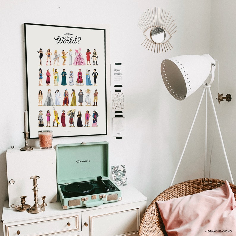 May include: A white wall with a framed poster that says "Who Run the World?" with a grid of colorful illustrations of women. A white lamp with a white shade is on a tripod stand. A green record player is on a white dresser with a gold candle holder. A wicker chair with a pink pillow is in the corner.