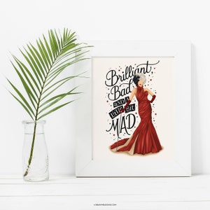 May include: A white framed print with a black and white illustration of a woman in a red dress. The text on the print reads "Brilliant, Bad and a little bit MAD".